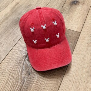 May include: Red baseball cap with white pearl accents arranged in Mickey Mouse shapes. The cap has a curved brim and a fabric-covered button on top. The hat is made of a soft, faded fabric.