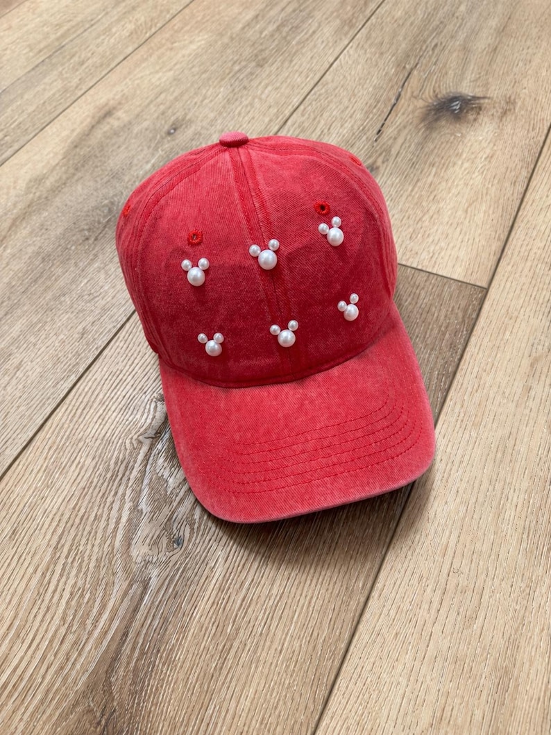 May include: Red baseball cap with white pearl accents arranged in Mickey Mouse shapes. The cap has a curved brim and a fabric-covered button on top. The hat is made of a soft, faded fabric.