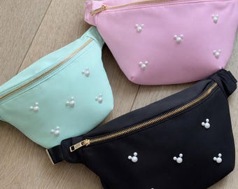 May include: Three fanny packs in pastel colors: mint green, light pink, and black. Each pack has a gold zipper and is adorned with small, white pearl embellishments arranged in a Mickey Mouse head shape. The bags are lying on a light wood surface.