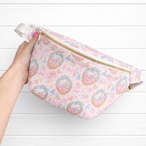 May include: Pink fanny pack with a repeating pattern of castles, bows, and Eiffel Towers. The bag has a gold zipper and a white strap. The overall design is whimsical and feminine.