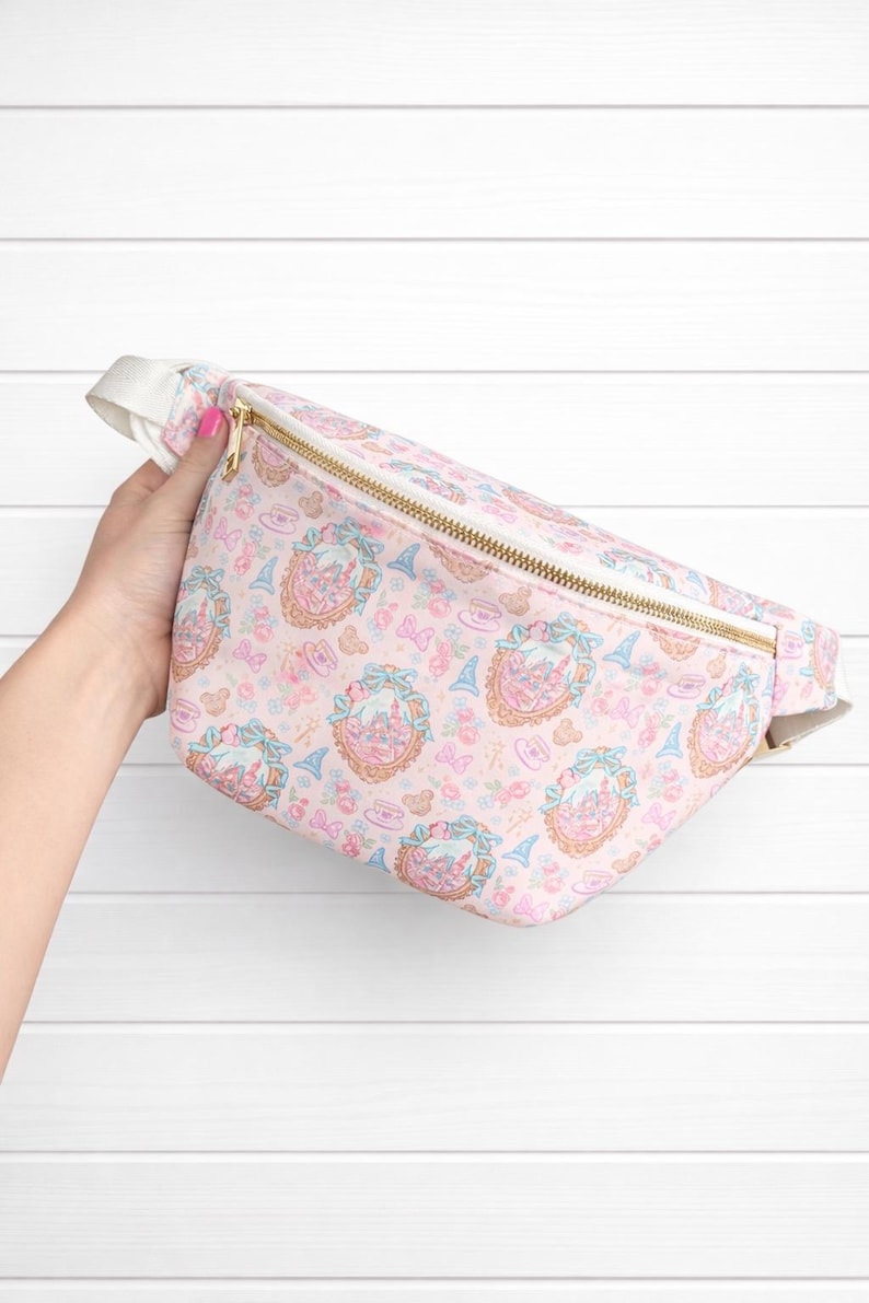 May include: Pink fanny pack with a repeating pattern of castles, bows, and Eiffel Towers. The bag has a gold zipper and a white strap. The overall design is whimsical and feminine.