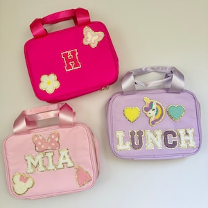 Könnte beinhalten: Drei Lunchboxen mit verschiedenen Designs. Die erste Tasche ist pink mit einem Schmetterling, einer Blume und dem Buchstaben "H" darauf. Die zweite Tasche ist lila mit einem Einhorn, zwei Herzen und dem Wort "LUNCH" darauf. Die dritte Tasche ist pink mit einer Schleife, dem Namen "MIA" und einem Herzen darauf.
