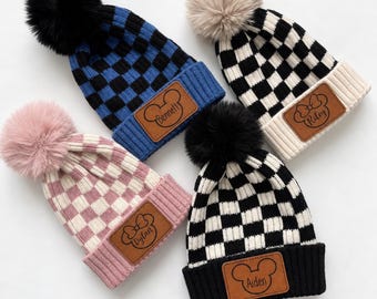 May include: Four knit beanies with pom-poms, each featuring a checkered pattern and a leather-like patch. The patches display names Bennett, Riley, Dylan, and Aiden alongside mouse-ear or bow outlines. Colors include blue, black, pink, and cream.