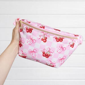 May include: Pink fanny pack with a gold zipper, featuring a repeating pattern of red cherries and pink bows on a white and pink checkered background. The bag is held up against a white background.