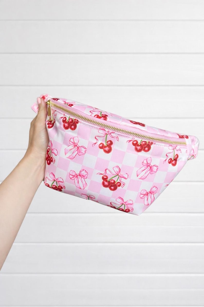 May include: Pink fanny pack with a gold zipper, featuring a repeating pattern of red cherries and pink bows on a white and pink checkered background. The bag is held up against a white background.
