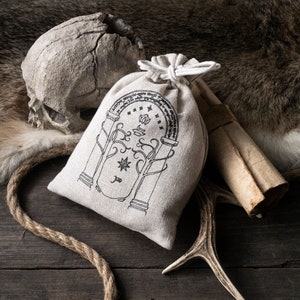 Könnte beinhalten: Ein weißer Stoffbeutel mit einem schwarzen Design eines Torbogens mit einem Stern und dem Text "The One Ring" auf einer Holzoberfläche mit einem Schädel, Geweih und einer aufgerollten Schriftrolle.