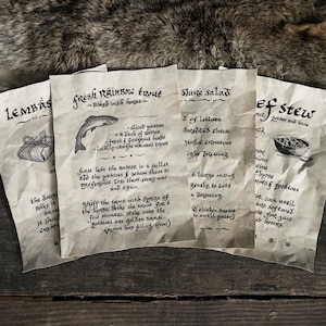 Op de afbeelding: Vier vellen verouderd papier met handgeschreven recepten, waaronder "Fresh Rainbow Trout" en "Beef Stew", zijn gerangschikt op een rustiek houten oppervlak. De tekst is in een gestileerd lettertype en illustraties van voedselitems begeleiden de tekst. De achtergrond omvat bont.