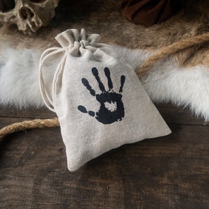 May include: A small, beige drawstring bag with a black handprint design. The bag is made of a natural fabric and has a simple, rustic look.