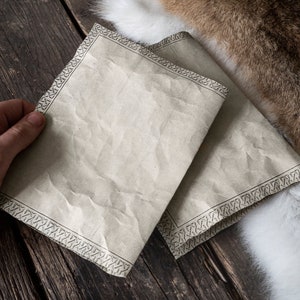 Może przedstawiać: Dwa arkusze papieru pergaminowego z ozdobną ramką. Papier jest lekko pomarszczony i ma vintage'owy wygląd. Papier leży na drewnianej powierzchni.