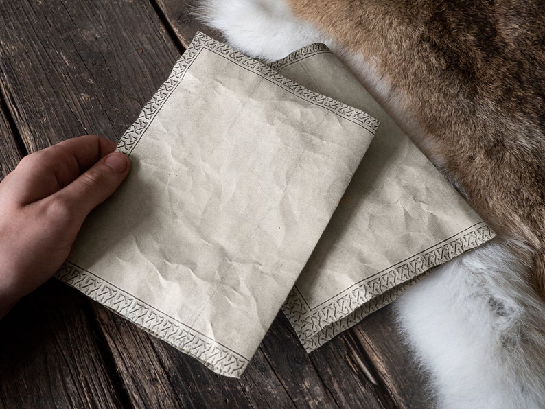 Może przedstawiać: Dwa arkusze papieru pergaminowego z ozdobną ramką. Papier jest lekko pomarszczony i ma vintage'owy wygląd. Papier leży na drewnianej powierzchni.