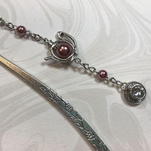 May include: Silver-toned metal bookmark with a decorative tea set charm. The charm features a teapot with a large, round, mauve bead and a teacup with a clear gem. The bookmark has a floral design along the edge and a curved top.