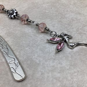 May include: A silver bookmark with an embossed floral design. The bookmark is connected to a chain with pink beads and a silver charm of a reclining fairy with pink gemstone wings. A unique accessory for book lovers.