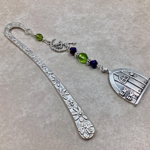 May include: A silver bookmark with a floral design and a charm. The charm features a fairy, green and purple beads, and a door with two figures. The bookmark is made of metal and is designed for books.