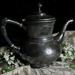 May include: Antique silver-plated teapot with a lid and ornate handle. The teapot features a detailed floral and scroll design, with a spout and a decorative handle. The surface shows signs of age and use, adding to its vintage charm.