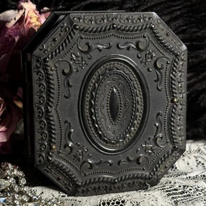May include: An octagonal, antique black box with intricate embossed floral and geometric patterns. The box features a central oval design and is embellished with small, gold-colored studs. The overall aesthetic is vintage and ornate.