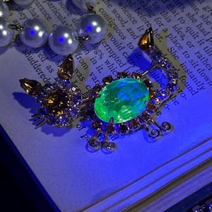 May include: A gold-toned scorpion brooch with a large, oval, green gemstone at its center. The scorpion's body is adorned with smaller amber and clear crystals. A pearl necklace is partially visible in the background.