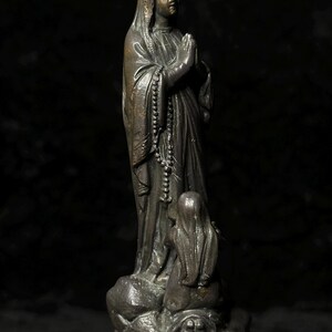 May include: A bronze statue of a robed figure with hands clasped in prayer, wearing a head covering and a rosary. A smaller figure kneels at the base. The statue has a dark, aged appearance.
