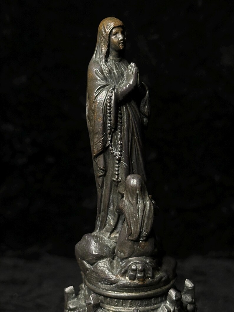 May include: A bronze statue of a robed figure with hands clasped in prayer, wearing a head covering and a rosary. A smaller figure kneels at the base. The statue has a dark, aged appearance.