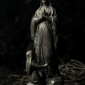 May include: A bronze statue of a religious figure with hands clasped in prayer, wearing a head covering and draped robes. A smaller figure kneels at the base, holding a staff. The statue is set against a dark background.