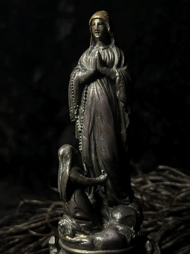 May include: A bronze statue of a religious figure with hands clasped in prayer, wearing a head covering and draped robes. A smaller figure kneels at the base, holding a staff. The statue is set against a dark background.