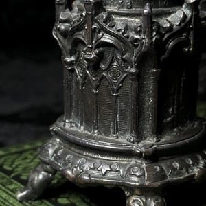 May include: Close-up of a detailed, antique-style metal object with ornate gothic architectural elements. The object has a dark, aged finish and is supported by decorative feet. The design includes arched windows and intricate carvings.