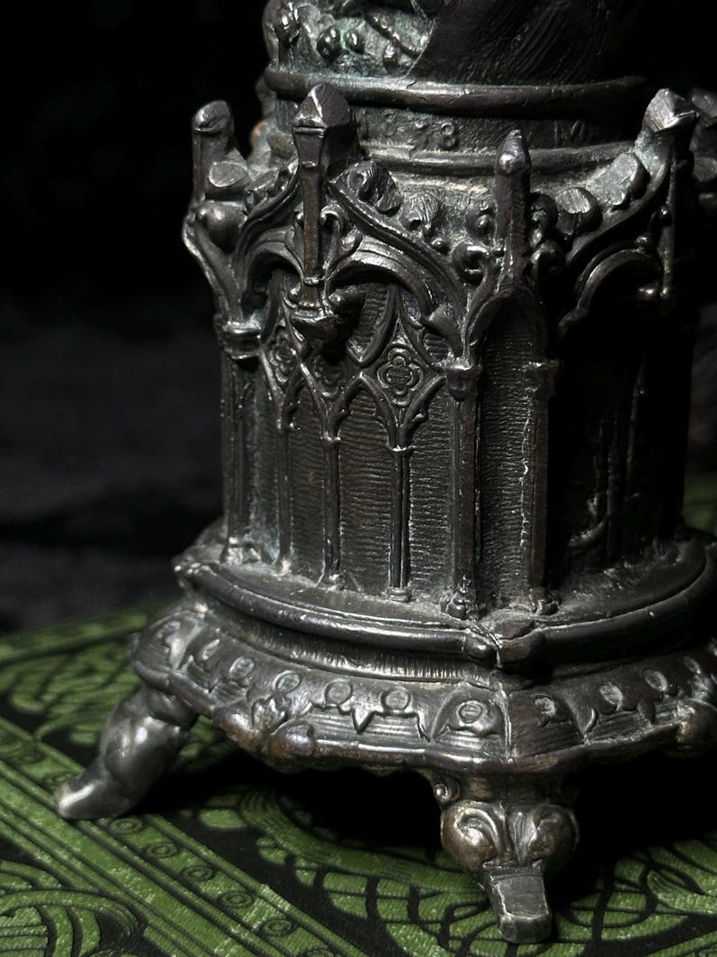 May include: Close-up of a detailed, antique-style metal object with ornate gothic architectural elements. The object has a dark, aged finish and is supported by decorative feet. The design includes arched windows and intricate carvings.