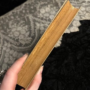 Peut inclure: Un livre ancien avec des pages jaunies et une tranche de couleur claire. La tranche du livre r&eacute;v&egrave;le le papier vieilli. Le livre est tenu en biais, soulignant son &eacute;tat usag&eacute; et son esth&eacute;tique vintage.
