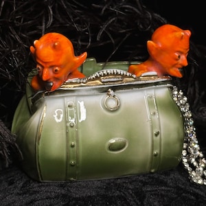 May include: A green ceramic purse with gold trim and two orange devil figurines peeking out. The purse has a handle and decorative rivets. A sparkling silver necklace is draped on the side.