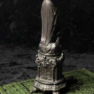 May include: A bronze-colored statue of a religious figure, possibly the Virgin Mary, stands on a detailed pedestal. The statue is set on a green book with an ornate design. The statue's robe has intricate detailing.
