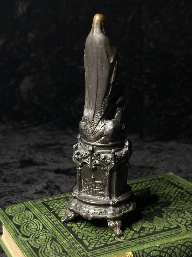 May include: A bronze-colored statue of a religious figure, possibly the Virgin Mary, stands on a detailed pedestal. The statue is set on a green book with an ornate design. The statue's robe has intricate detailing.
