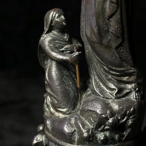May include: A bronze statue depicting a kneeling figure looking up at a larger figure. The smaller figure holds a staff. The statue has intricate details and is set against a dark background.