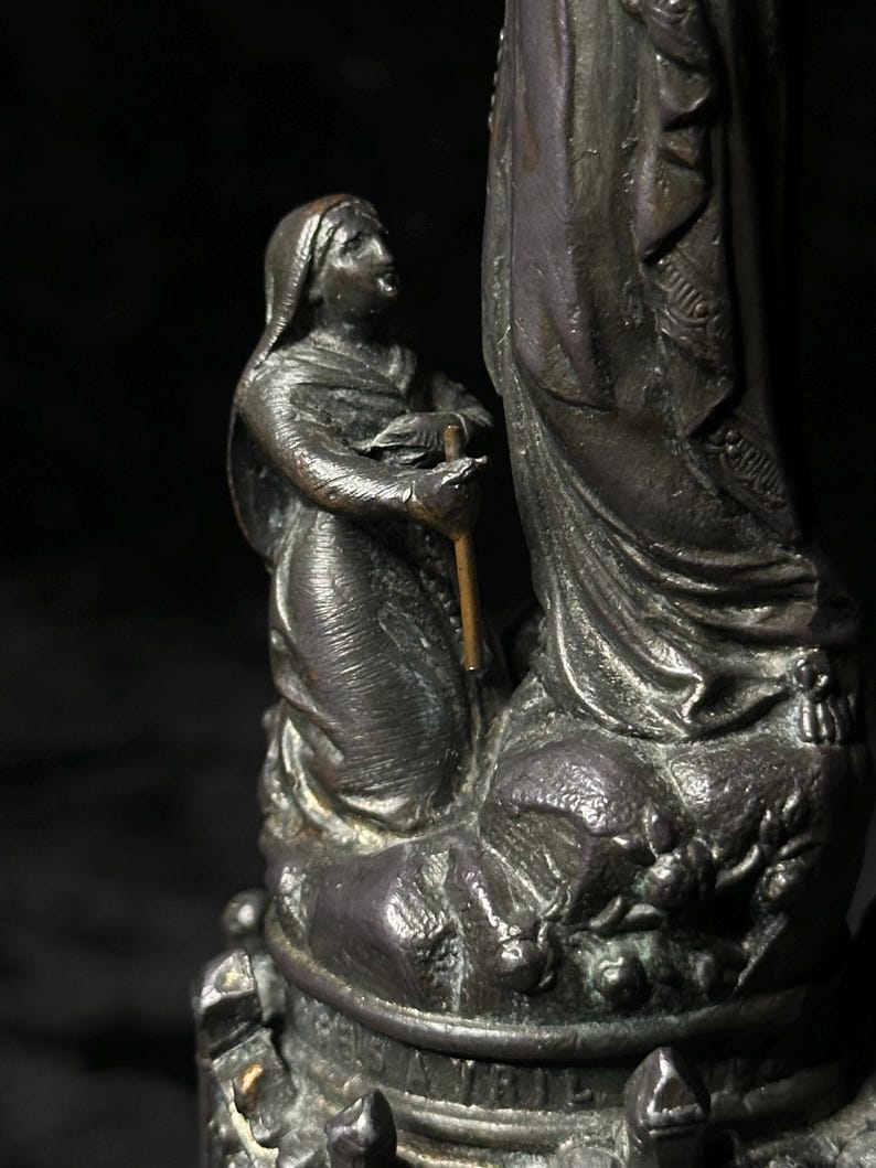 May include: A bronze statue depicting a kneeling figure looking up at a larger figure. The smaller figure holds a staff. The statue has intricate details and is set against a dark background.