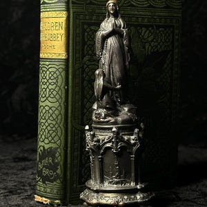 May include: A dark metal statue of a woman in prayer, with a child at her feet, atop an ornate base. Behind it is a vintage green book with gold lettering that reads "CHILDREN OF THE ABBEY" and "BUTLER BROS."