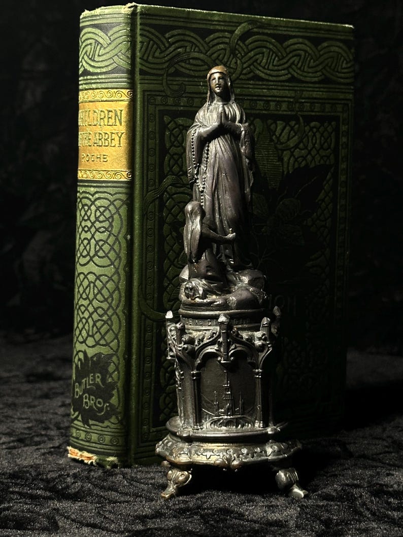 May include: A dark metal statue of a woman in prayer, with a child at her feet, atop an ornate base. Behind it is a vintage green book with gold lettering that reads "CHILDREN OF THE ABBEY" and "BUTLER BROS."