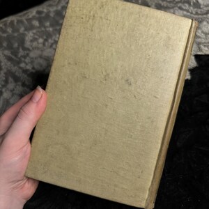Peut inclure: Un livre vintage avec une couverture beige clair, tenu par une main. La couverture montre des signes de vieillissement avec des taches plus fonc&eacute;es et un aspect l&eacute;g&egrave;rement us&eacute;. Le dos est d'une teinte plus fonc&eacute;e de beige. Le fond est un tissu sombre et textur&eacute;.