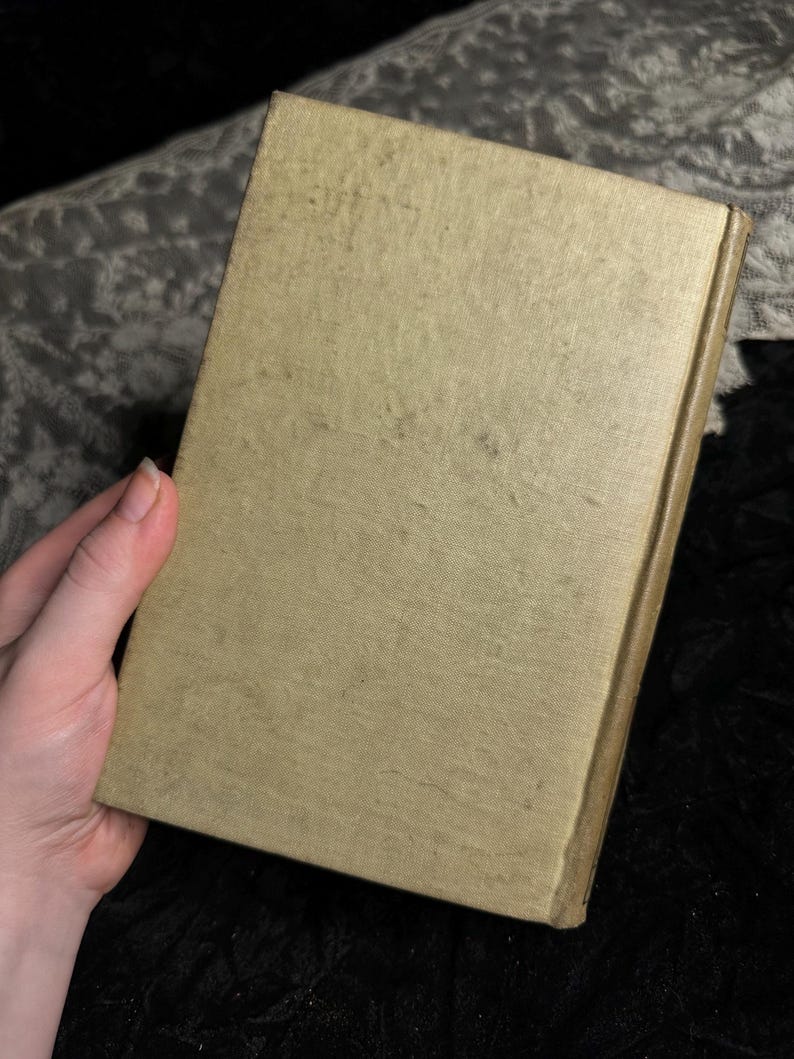 Peut inclure: Un livre vintage avec une couverture beige clair, tenu par une main. La couverture montre des signes de vieillissement avec des taches plus fonc&eacute;es et un aspect l&eacute;g&egrave;rement us&eacute;. Le dos est d'une teinte plus fonc&eacute;e de beige. Le fond est un tissu sombre et textur&eacute;.