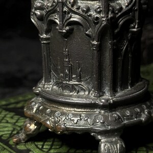 May include: A detailed, antique-style metal object with a dark, aged finish. The object features ornate architectural details, including arched designs and a depiction of a building. It rests on decorative feet and is likely a decorative item.