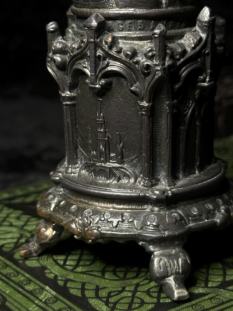 May include: A detailed, antique-style metal object with a dark, aged finish. The object features ornate architectural details, including arched designs and a depiction of a building. It rests on decorative feet and is likely a decorative item.