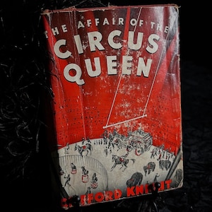 May include: A vintage book cover with a red background and the title "The Affair of the Circus Queen" in white lettering. The cover features a black and white illustration of a circus ring with performers and animals.