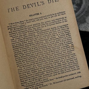 Peut inclure: Page d'un livre ouvert avec le titre "THE DEVIL'S DIE" en haut. Le texte est encre noire sur du papier vieilli, blanc cass&eacute;. Le premier chapitre commence par une description d'une sc&egrave;ne de gare.