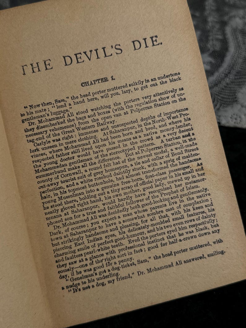 Peut inclure: Page d'un livre ouvert avec le titre "THE DEVIL'S DIE" en haut. Le texte est encre noire sur du papier vieilli, blanc cass&eacute;. Le premier chapitre commence par une description d'une sc&egrave;ne de gare.