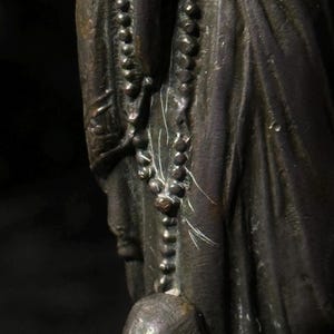 May include: A bronze statue depicting a figure wearing draped clothing and a beaded necklace. The statue has a dark patina and shows signs of age and wear. The figure's face is partially visible.