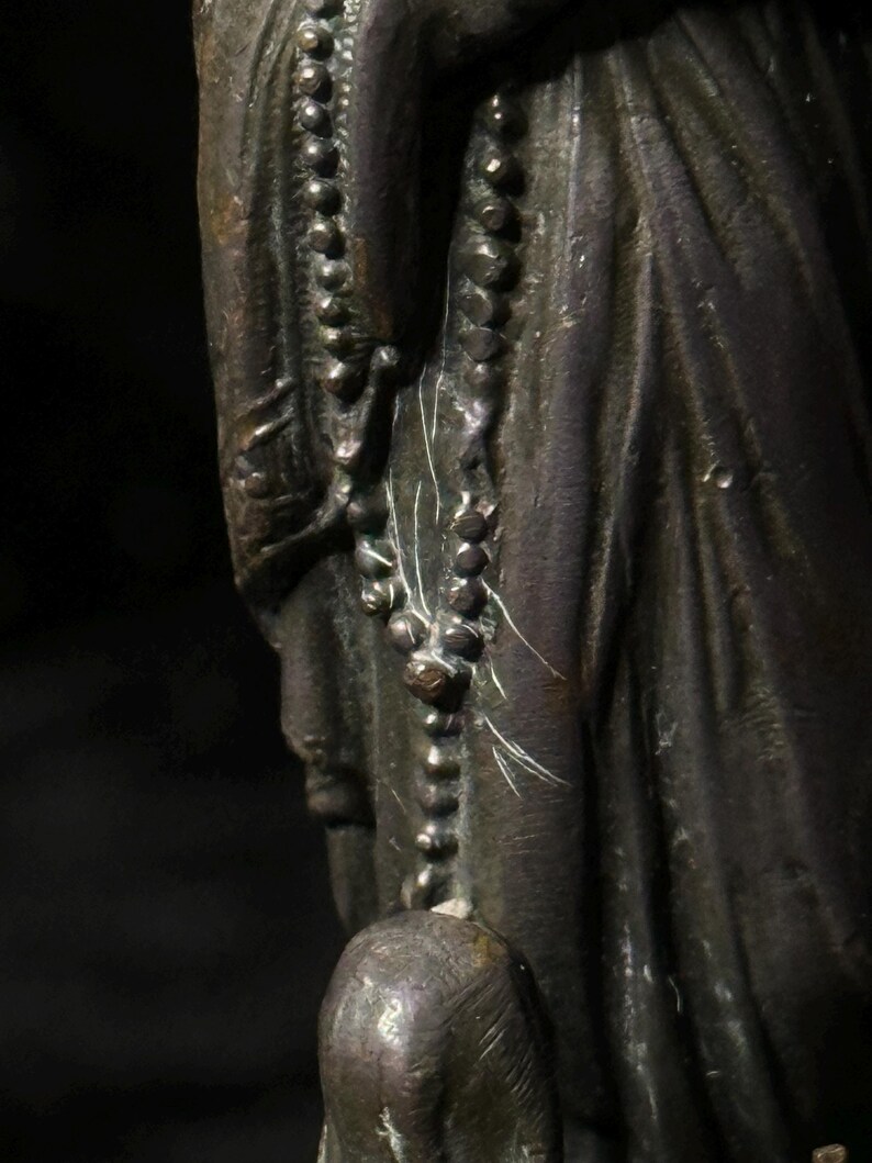 May include: A bronze statue depicting a figure wearing draped clothing and a beaded necklace. The statue has a dark patina and shows signs of age and wear. The figure's face is partially visible.