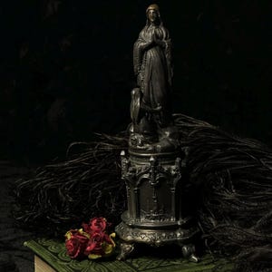 May include: A dark, antique-style statue of a woman in prayer, with an angel at the base, atop a decorative stand. The statue is silver-toned, with intricate detailing. Dried red roses and black feathers are in the foreground, resting on a green book.