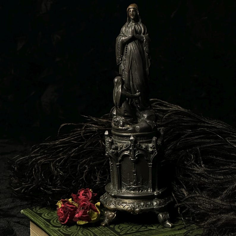 May include: A dark, antique-style statue of a woman in prayer, with an angel at the base, atop a decorative stand. The statue is silver-toned, with intricate detailing. Dried red roses and black feathers are in the foreground, resting on a green book.