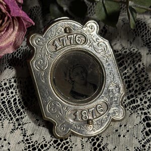 May include: An antique silver-toned locket with an oval portrait inside. The locket is rectangular with ornate scrollwork and the dates "1776" and "1876" engraved on it. The locket is resting on a white lace surface.