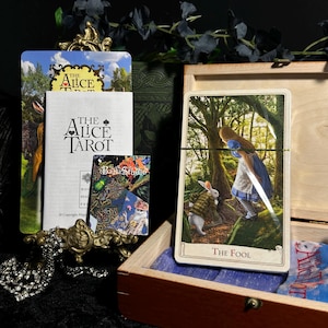 May include: An open wooden box contains an "Alice Tarot" deck, a guidebook, and a small card with a colorful design. The "Fool" tarot card is visible, depicting Alice and the White Rabbit. A decorative metal piece and a necklace are in the foreground.