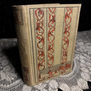 May include: A vintage book with a tan cover and decorative orange and black floral patterns. The spine features the title "THE DEVIL'S BIBLE" in gold lettering. The book is displayed on a white lace surface.