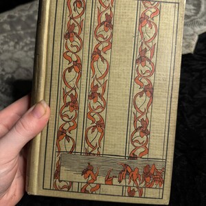 Peut inclure: Un livre ancien avec une couverture dor&eacute;e orn&eacute;e de motifs floraux orange verticaux. La couverture pr&eacute;sente un design rectangulaire avec trois colonnes de fleurs et de feuillages stylis&eacute;s. Le dos du livre est visible et les pages sont vieillies.