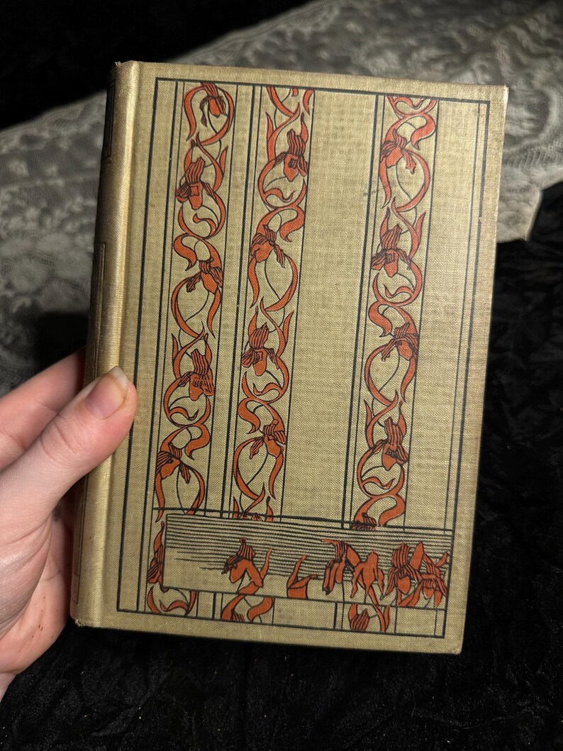 Peut inclure: Un livre ancien avec une couverture dor&eacute;e orn&eacute;e de motifs floraux orange verticaux. La couverture pr&eacute;sente un design rectangulaire avec trois colonnes de fleurs et de feuillages stylis&eacute;s. Le dos du livre est visible et les pages sont vieillies.
